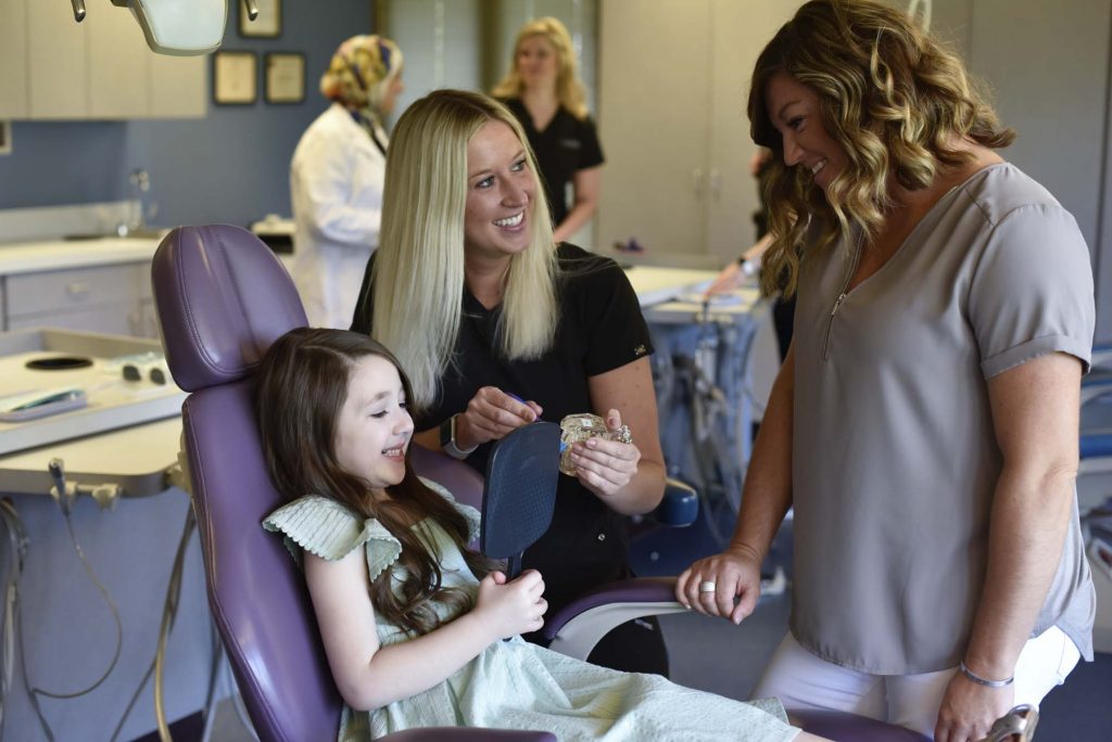 a smiling child sits in a dental chair holding a mirror while two female employees smile at her