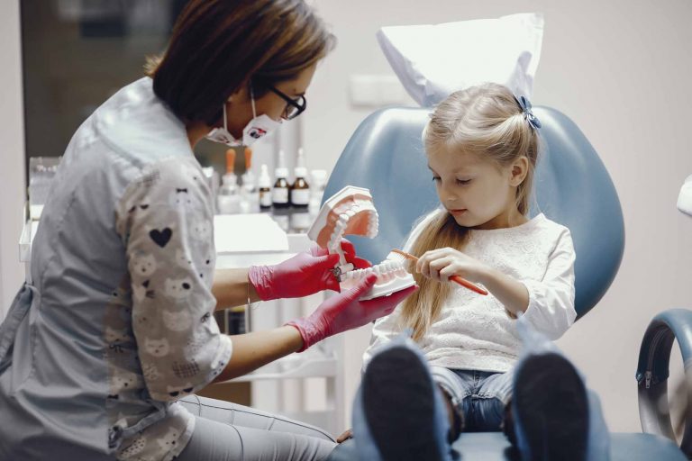 child sitting in dental chair brushing a tooth model held by a dental hygienist