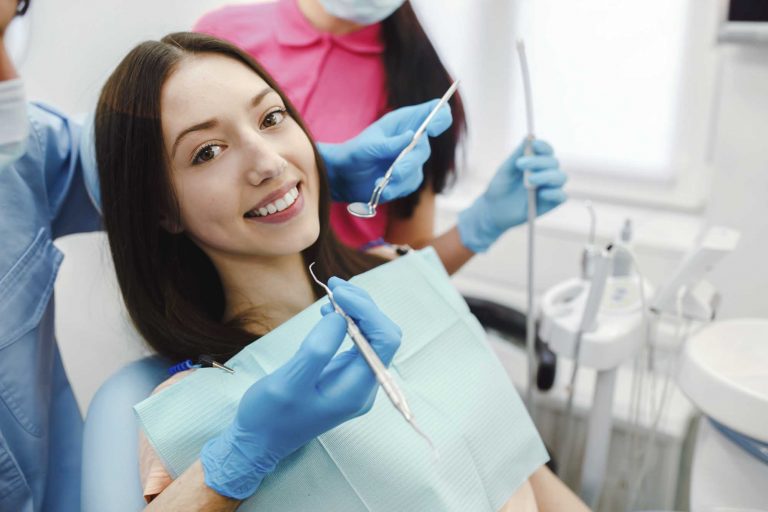 smiling woman in dental chair while a hygienist holds dental tools near her