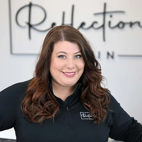 Headshot of April wearing a black jacket with 'Dental Reflections Dublin' logo, standing against a blurred background featuring the same logo.