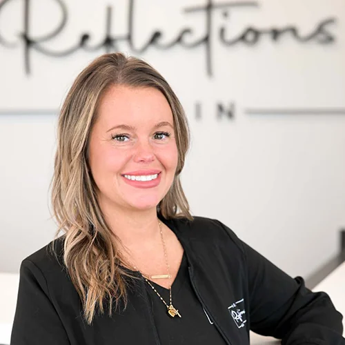 Headshot of Chassiti wearing a black jacket with 'Dental Reflections Dublin' logo, standing against a blurred background featuring the same logo.