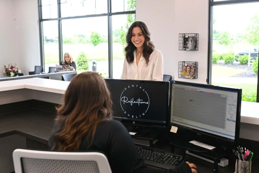 Reception area of a dental office with two women at the desk; one seated using a computer, and the other standing. Background includes large windows showing greenery, seating area, and wall decorations.